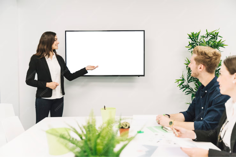 Woman presenting to two colleagues in a meeting room, pointing to a blank screen.